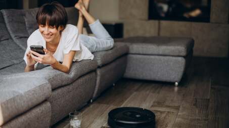 woman lying on couch, using her phone, while a robot vacuum cleans the floor 